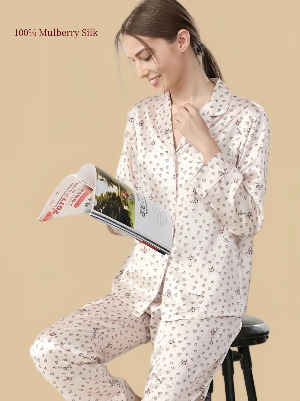 Model reading in Silkgala Pale Blush Pink silk pajamas with small cat illustrations, outlined hearts, long sleeves and matching relaxed pants.