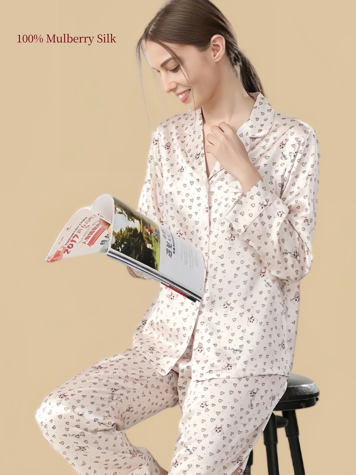 Model reading in Silkgala Pale Blush Pink silk pajamas with small cat illustrations, outlined hearts, long sleeves and matching relaxed pants.