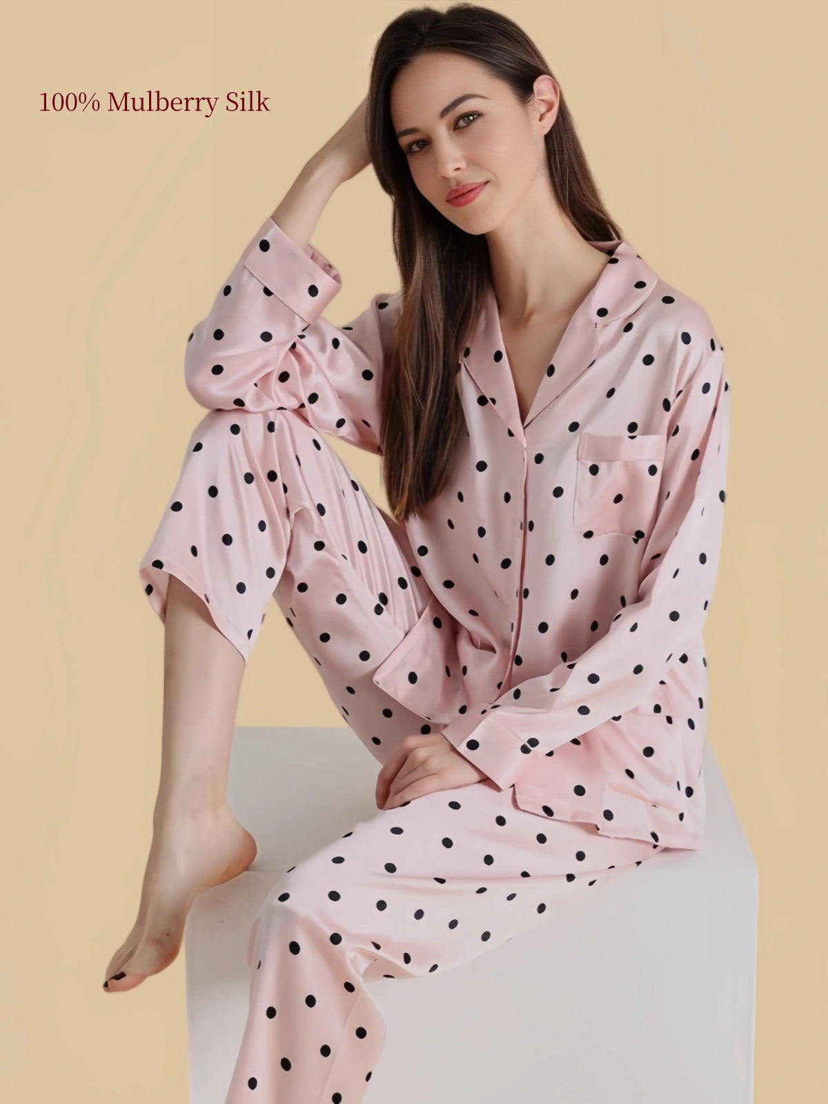 Model seated in Silkgala blush pink silk pajamas with black polka dots, showing the button-front shirt, chest pocket and relaxed pants.